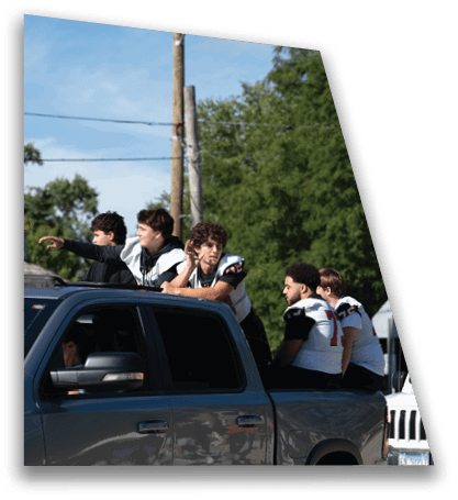 Several people wearing sports jerseys are sitting and leaning on the back of a dark pickup truck parked on a street with trees and power lines in the background. Description generated by AI