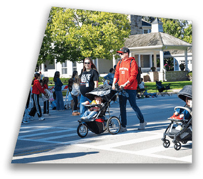 Adults wearing casual clothing are pushing children in strollers while crossing a street in a sunny neighborhood with houses and trees in the background. Description generated by AI