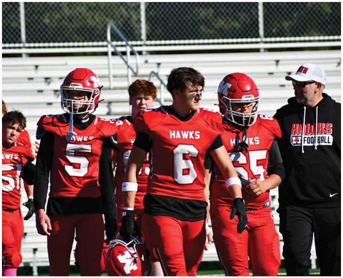 People wearing red football uniforms with “HAWKS" on the jerseys are standing on a field, with one person holding a helmet and another wearing a black hoodie with "HAWKS FOOTBALL" on it. Description generated by AI