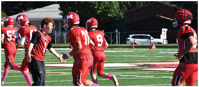 People wearing red football uniforms are on a green field with white yard lines, with one person in a black and red outfit reaching out to a player. Description generated by AI