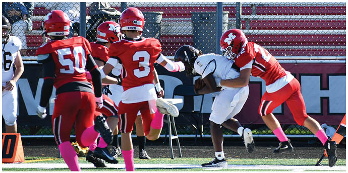 Several people in football uniforms are on a field near a chain-link fence with spectators in the background, with some players wearing red and others in white. Description generated by AI