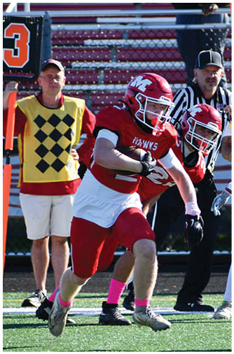 Two people in football uniforms are positioned on a field with one wearing a red jersey and helmet facing the other in a white jersey and helmet, while a person in casual clothing stands in the background near a down marker. Description generated by AI