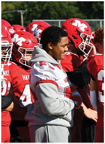 A group of people wearing red football uniforms and helmets stand together on a field with one person in a gray hoodie and white jersey beside them. Description generated by AI