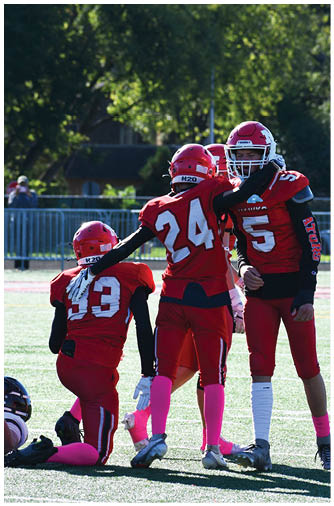 Two people wearing red football uniforms and helmets stand on a sunlit field with one person placing a hand on the other's shoulder. Description generated by AI