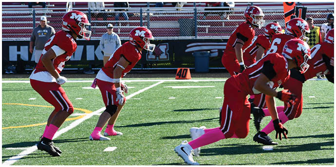 Several people wearing red football uniforms and helmets stand together on a field with a scoreboard and buildings in the background. Description generated by AI