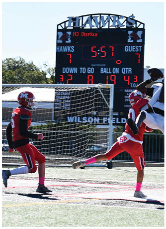 Two athletes in red and white uniforms are competing on a football field with one player leaping to catch the ball while the scoreboard in the background shows the home team leading. Description generated by AI