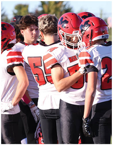 Two people wearing red helmets and white football jerseys with red numbers are standing close together on a field, with one person holding the other's shoulder. Description generated by AI