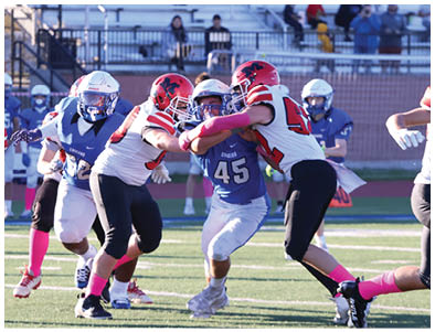 Players in red and white uniforms with pink socks are actively engaged in a football game against players in blue uniforms on a field with empty bleachers in the background. Description generated by AI
