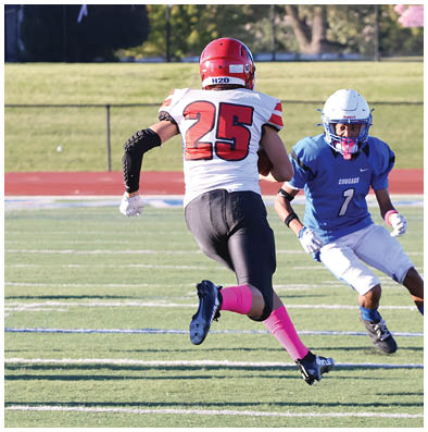 Two people in football uniforms are actively engaged on a field with one person in a blue jersey and white pants facing another in a white jersey with red and black accents and pink socks. Description generated by AI