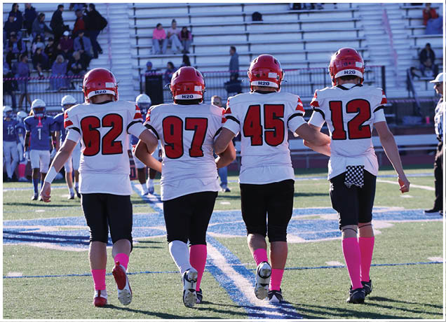Four football players wearing red helmets, white jerseys with black and red accents, black pants, and pink socks walk arm in arm on a football field toward the stands. Description generated by AI