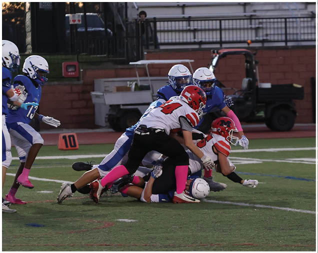 Several people in football uniforms are engaged in a tackle on a field with a few others standing nearby and empty bleachers in the background. Description generated by AI