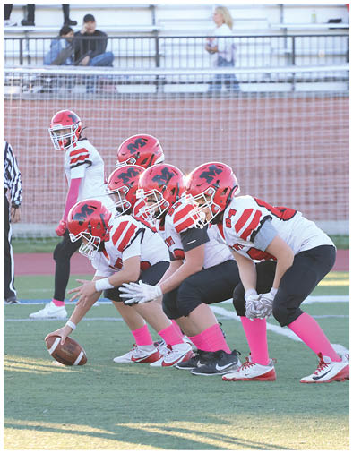 Five people in football uniforms with red helmets and pink socks are lined up on a field in a ready stance, preparing for a play. Description generated by AI
