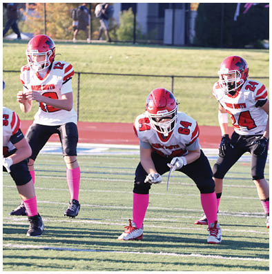 Three people wearing red helmets, white jerseys with red and black details, black pants, and pink socks are positioned on a football field, appearing ready for a play. Description generated by AI