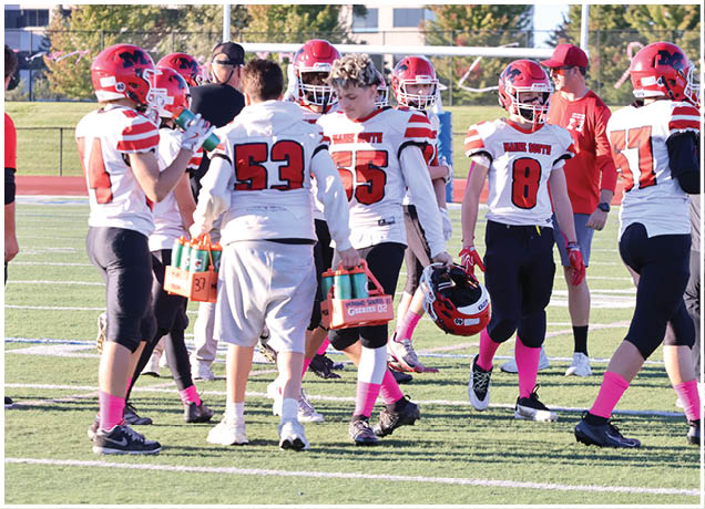 A group of people wearing football uniforms and helmets are gathered on a field, some holding water bottles and one holding a helmet. Description generated by AI