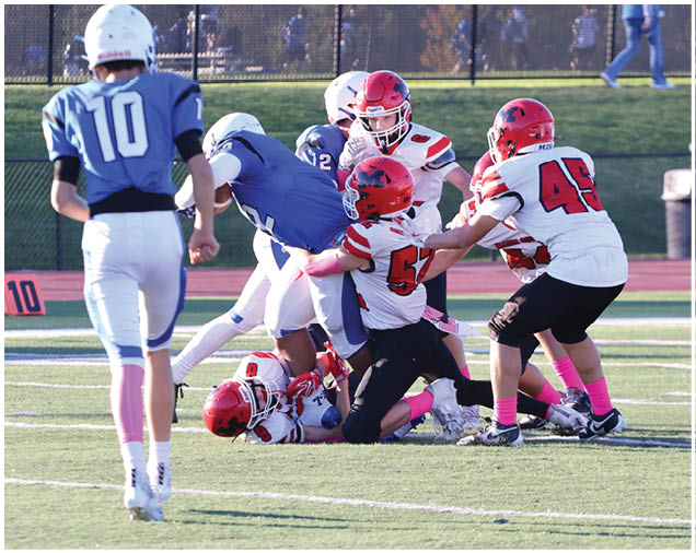 Several people wearing football uniforms are engaged in a play on a grassy field with some players tackling others while one person stands nearby. Description generated by AI