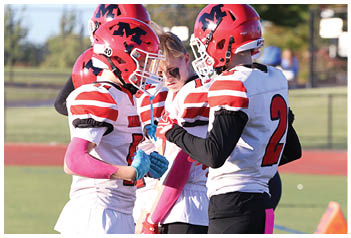 Three people wearing red and white football uniforms and helmets stand on a field while two others in similar uniforms stand in the background. Description generated by AI