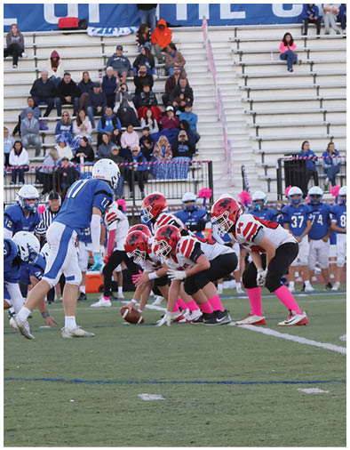 Four people wearing red helmets, white jerseys with red stripes, black pants, and pink socks are running on a football field with bleachers and spectators in the background. Description generated by AI