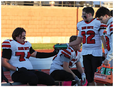 People wearing sports uniforms sit on a bench while one person hands a water bottle to another during a game or practice. Description generated by AI