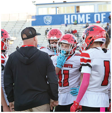 A group of people in football uniforms and helmets are gathered on a field with one person in a black hoodie and cap facing them. Description generated by AI
