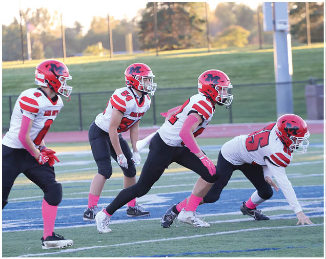 Four people wearing red helmets, white and red football jerseys, and pink accessories are positioned on a football field preparing for a play. Description generated by AI