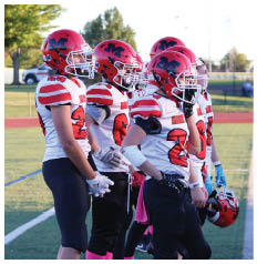 Four people wearing red helmets and white football jerseys with red stripes stand in a row on a sports field during daylight. Description generated by AI