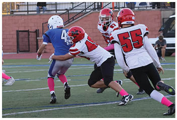 People wearing blue and red football uniforms are actively playing on a field with empty bleachers in the background during a game. Description generated by AI