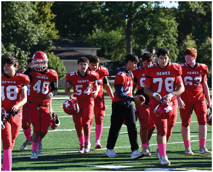 A group of people wearing red football uniforms with pink socks are walking together on a green football field with trees and houses in the background. Description generated by AI