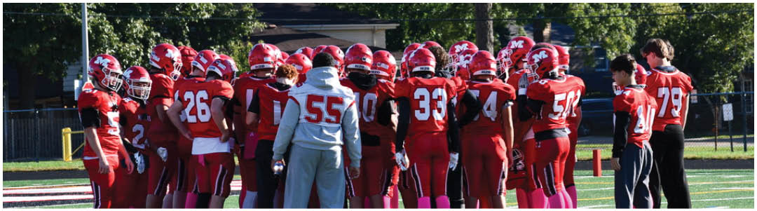 A group of people wearing red football uniforms and helmets are gathered closely together on a grassy field with trees and a building in the background. Description generated by AI
