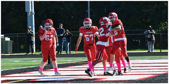 Five people in red football uniforms are on a field with one person holding a football while others gather around, and several people stand in the background near a fence. Description generated by AI