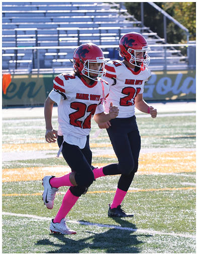 Two people wearing red helmets, white jerseys with “Maine South" text, black pants, and pink socks are running on a football field. Description generated by AI