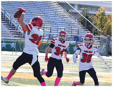 Four people wearing football uniforms and helmets are on a field with bleachers in the background, with three in white jerseys running toward one in a green jersey. Description generated by AI