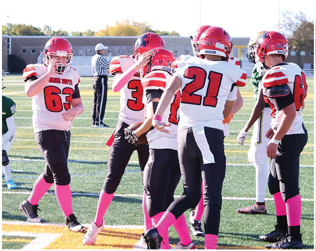 Several people wearing football uniforms with red helmets and pink socks are on a field with one person in a green uniform running behind them. Description generated by AI