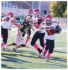 People wearing football uniforms with pink socks are actively playing on a sunlit field with one person running while holding the ball and others preparing to block or tackle. Description generated by AI