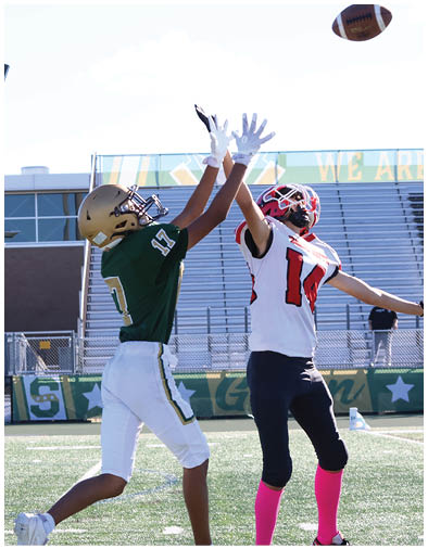 Two people in football uniforms with helmets and gloves are reaching up to catch a football on a field with bleachers in the background. Description generated by AI