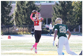 A person wearing a white and red sports uniform with pink socks is jumping to catch a football while two other players in green and white and white and red uniforms run nearby on a sunlit field. Description generated by AI
