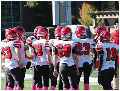 A group of people wearing red helmets and white sports jerseys with black pants are standing together on a sports field. Description generated by AI