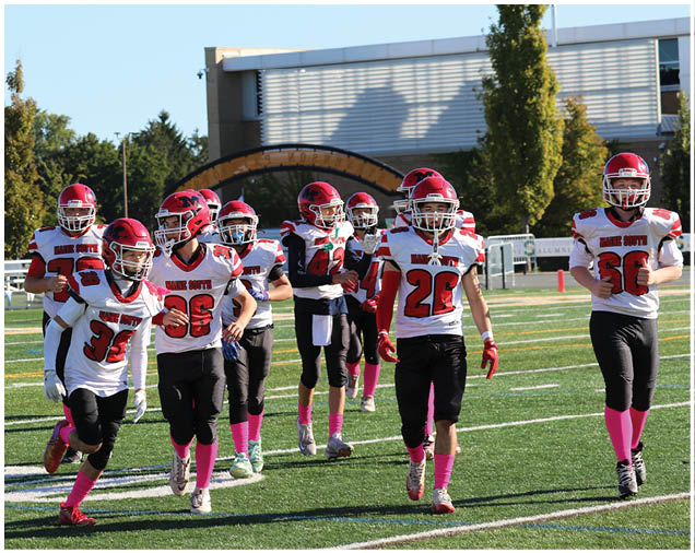 A group of people wearing red helmets, white jerseys, black pants, and pink socks are walking on a football field with trees and a building in the background. Description generated by AI