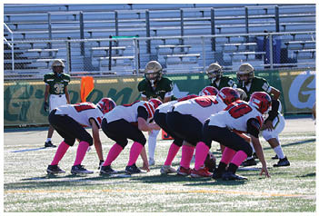 Several people in football uniforms with red helmets and pink socks are lined up on a field facing a person in a green uniform standing behind them. Description generated by AI