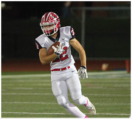 A person wearing a white and red football uniform is running on a field while holding a football, with another person in a blue and gray uniform partially visible in the foreground. Description generated by AI