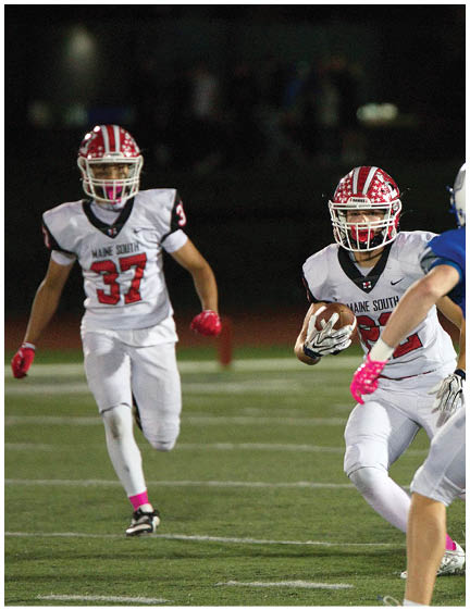 Two people in white football uniforms with red helmets are running on a field while one holds a football and another person in a blue uniform is approaching. Description generated by AI