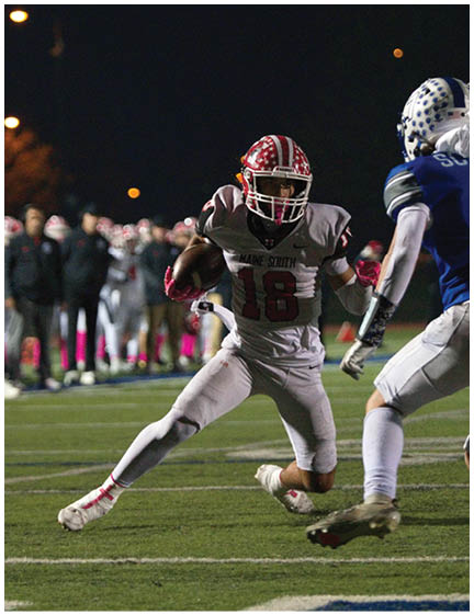 A person wearing white athletic shoes and socks with red stripes is running on a football field with a group of people and players in white uniforms standing in the background at night. Description generated by AI