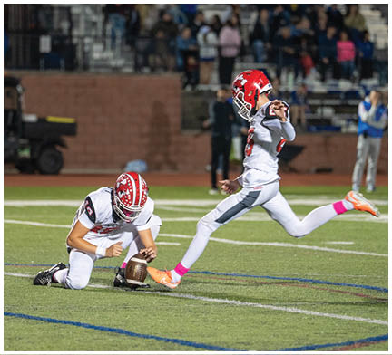 Two people in football uniforms are on a field with one holding the ball on the ground while the other kicks it, likely during a field goal attempt. Description generated by AI
