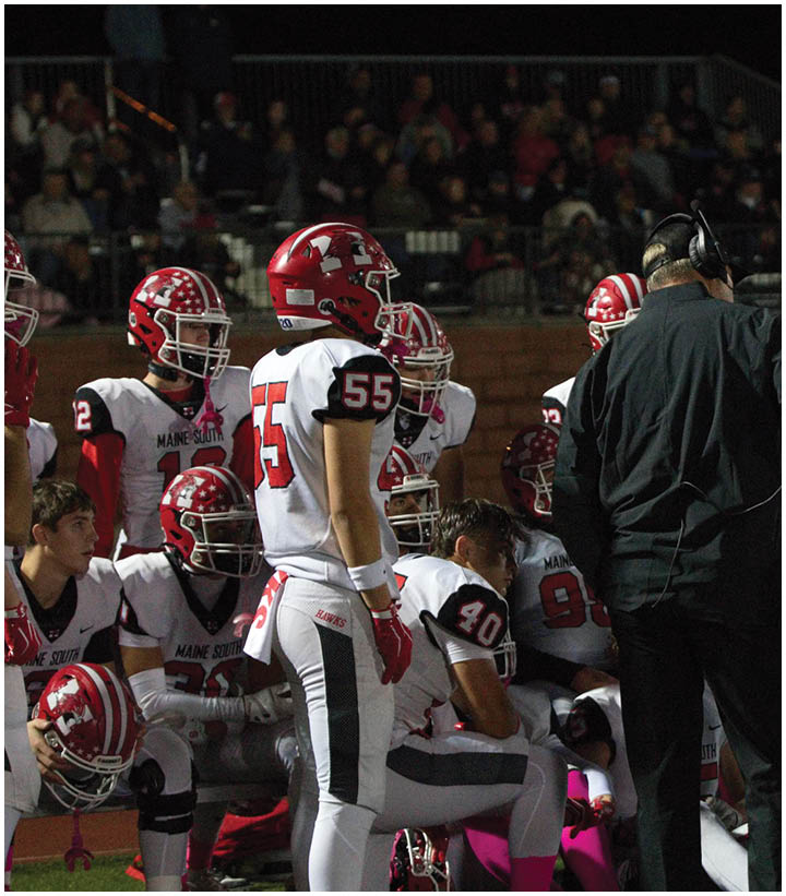 A group of football players in white and red uniforms are gathered around a coach wearing a headset who is standing next to a large monitor on the sidelines during a nighttime game. Description generated by AI