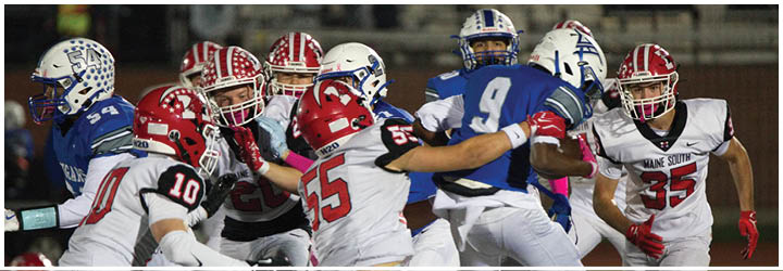 Several people in football uniforms are actively engaged in a play on a field, with one person holding the ball while others attempt to block or tackle. Description generated by AI