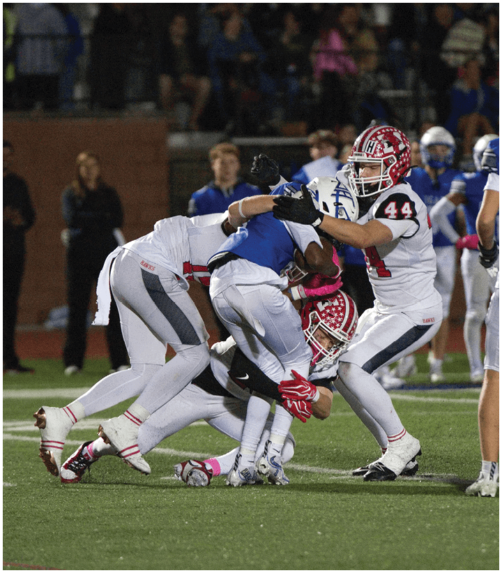 Three football players in white uniforms tackle a player in a blue uniform on a grassy field during a game with spectators in the background. Description generated by AI