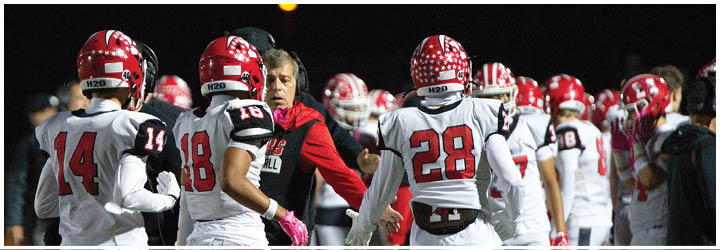 A group of people wearing red and white football helmets and jerseys are gathered around a person in a red and black jacket who appears to be giving instructions. Description generated by AI