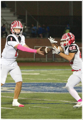 Two people in football uniforms are on a field with one handing off a football to the other during a game at night. Description generated by AI