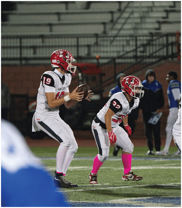 Two people in football uniforms with red helmets and pink socks are on a field, one holding a football in a ready-to-throw position while the other is crouched in a defensive stance. Description generated by AI