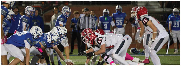 Two opposing American football teams in blue and white and red and white uniforms are lined up facing each other on a field with referees and other players in the background. Description generated by AI