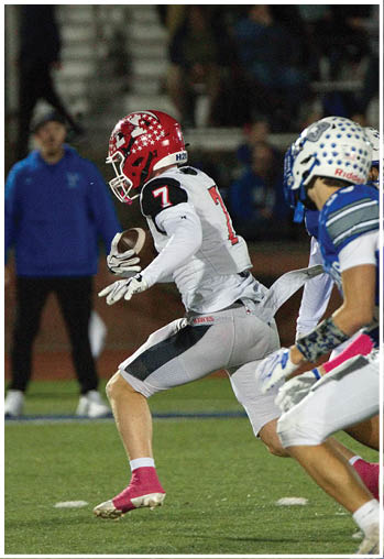 A person wearing a blue sports jersey with the number seven and a white helmet is running on a football field while two people stand in the background. Description generated by AI